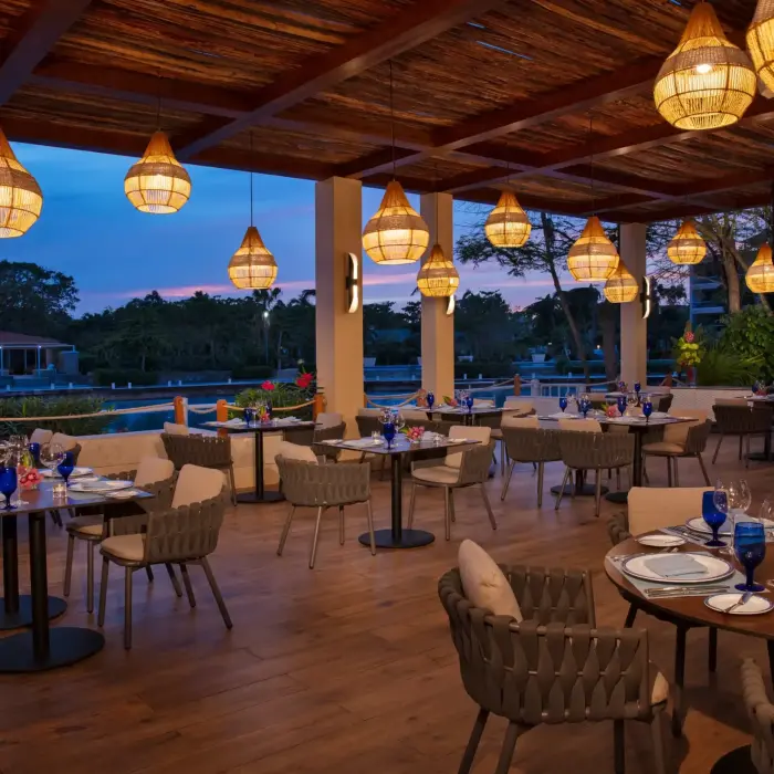 Open-air terrace restaurant with wicker lighting, wooden ceiling, and tranquil lagoon views at sunset.