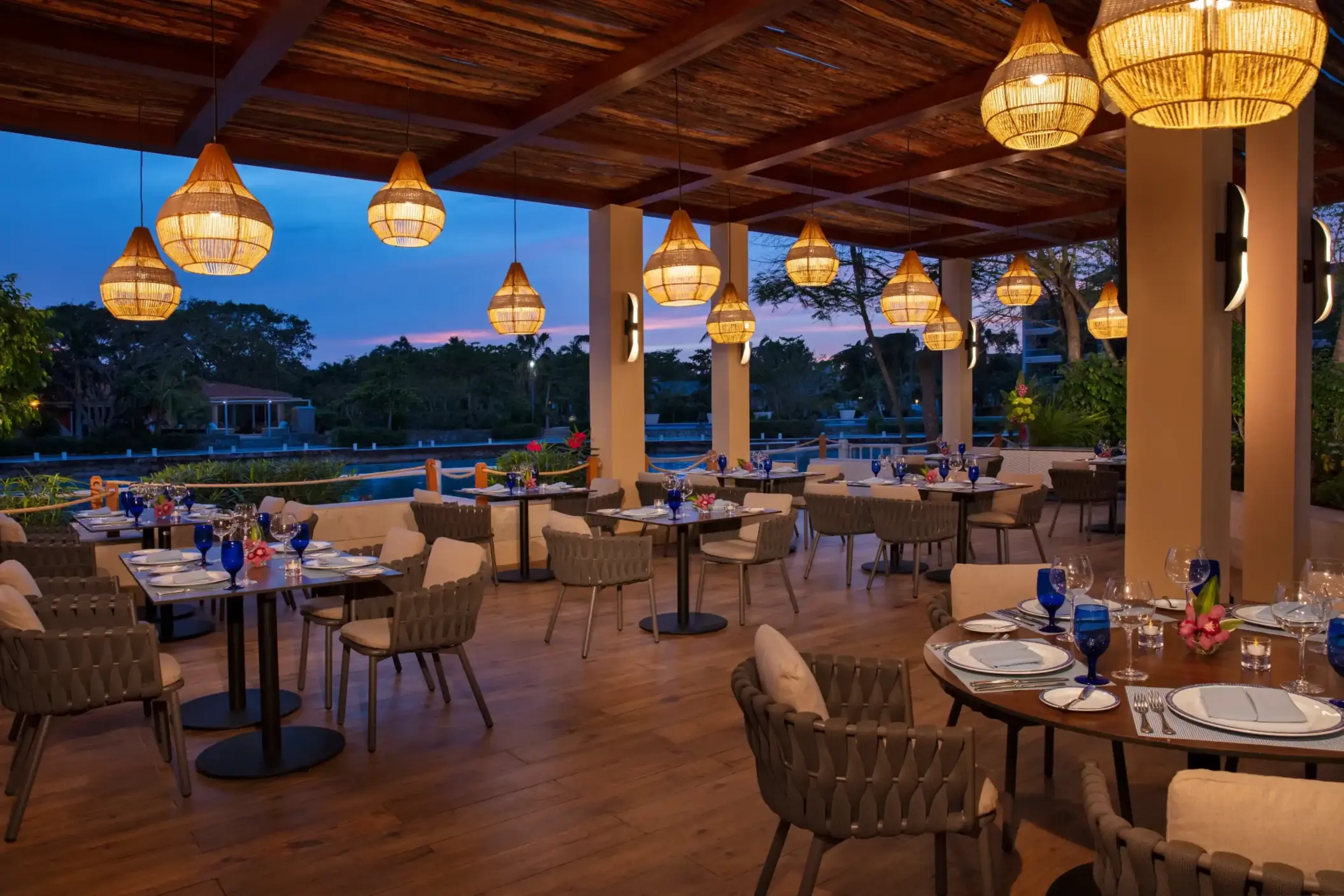 Open-air terrace restaurant with wicker lighting, wooden ceiling, and tranquil lagoon views at sunset.