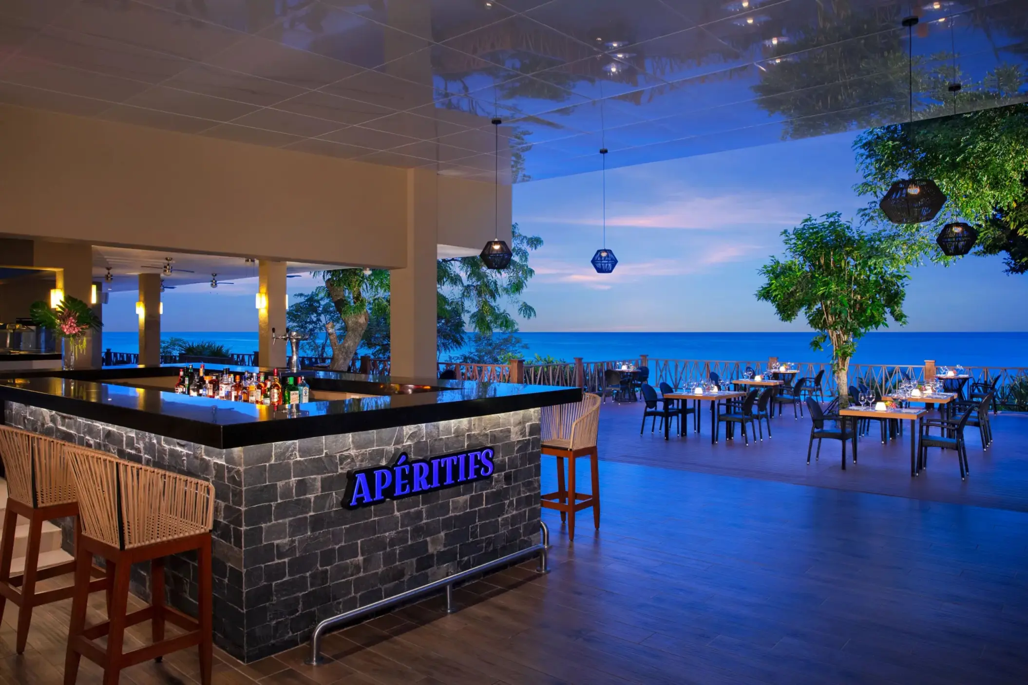 Elegant Aperitifs bar with oceanview terrace dining, glowing lights, and modern tropical decor at dusk.