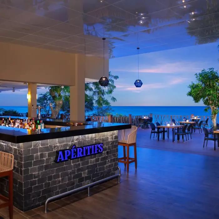 Elegant Aperitifs bar with oceanview terrace dining, glowing lights, and modern tropical decor at dusk.