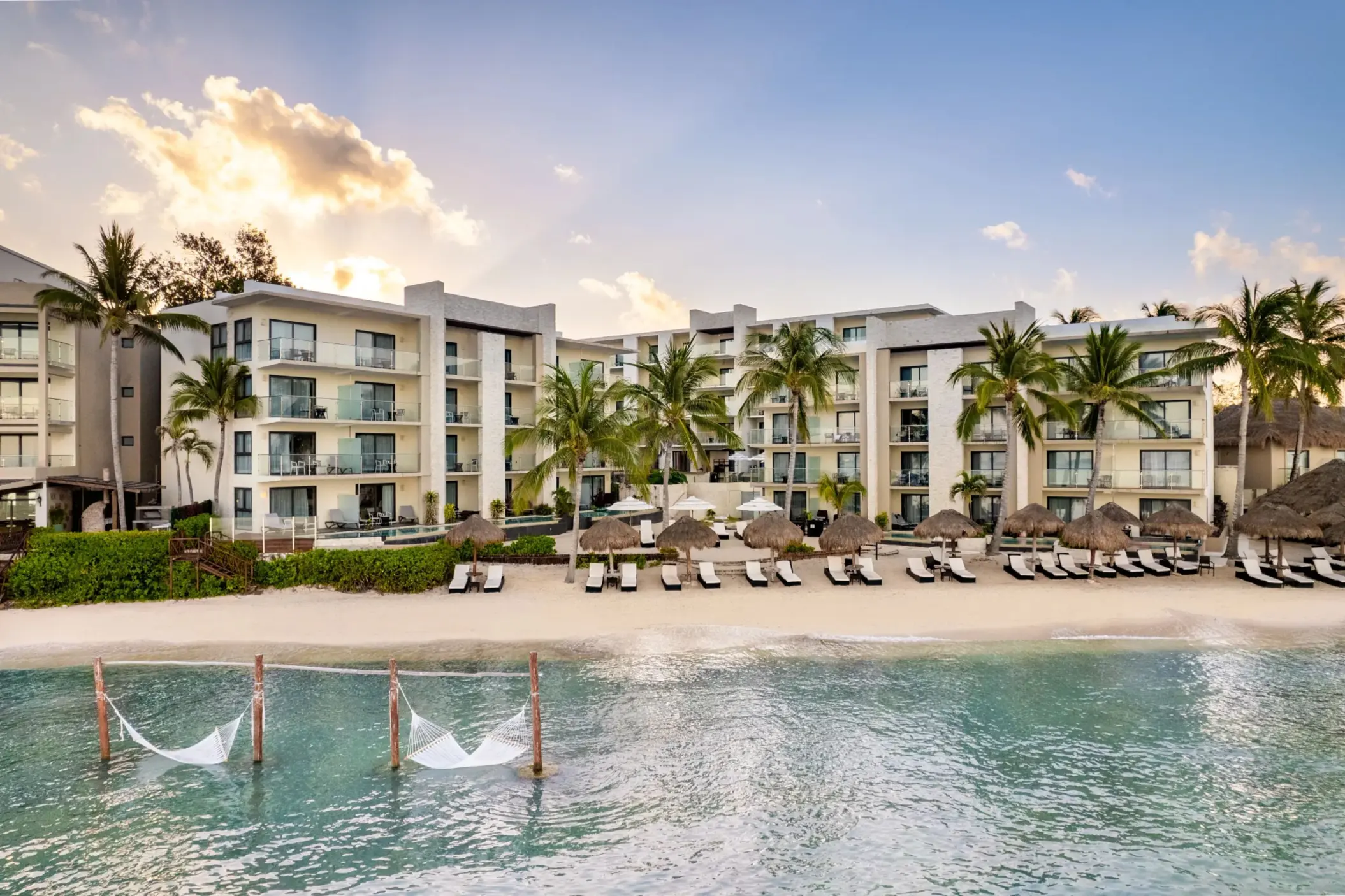 Ocean-facing resort with white sand beach, lounge chairs, and tranquil water framed by tropical trees.