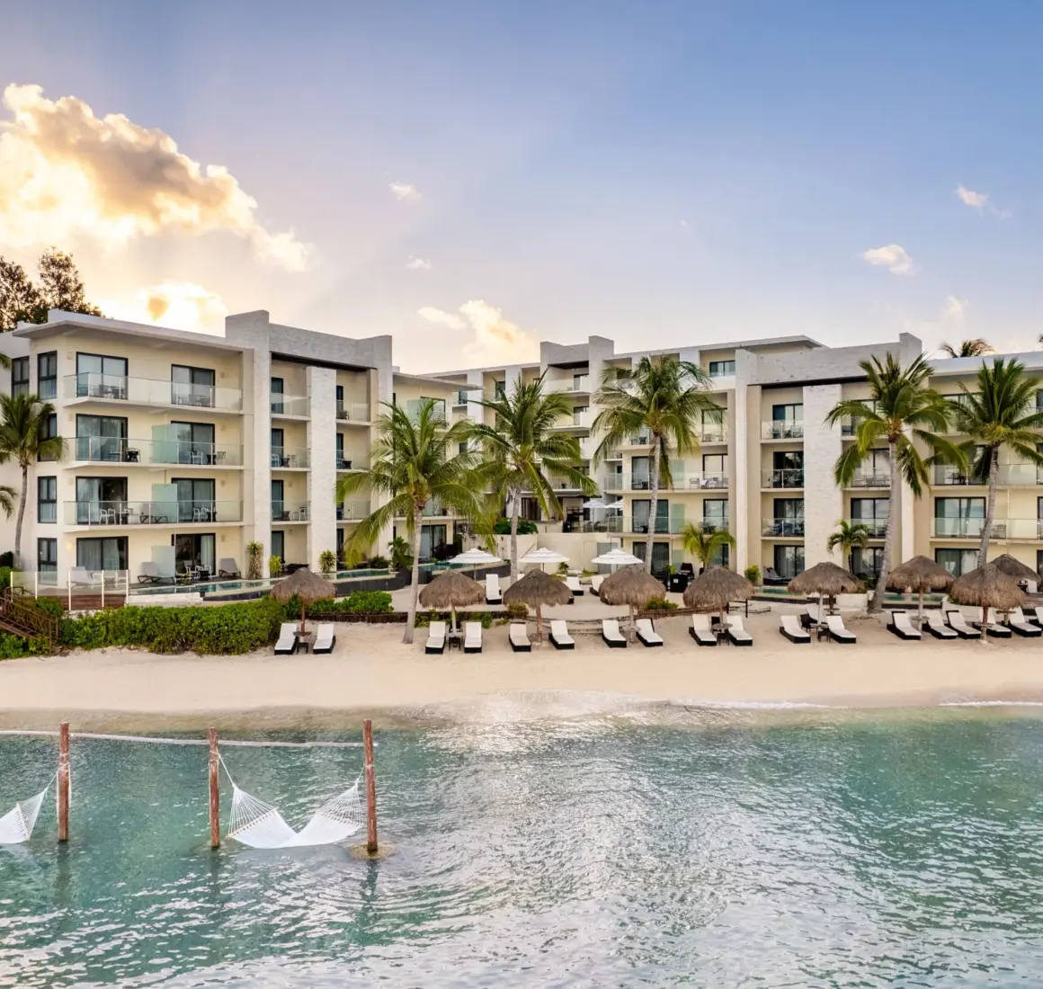 Ocean-facing resort with white sand beach, lounge chairs, and tranquil water framed by tropical trees.