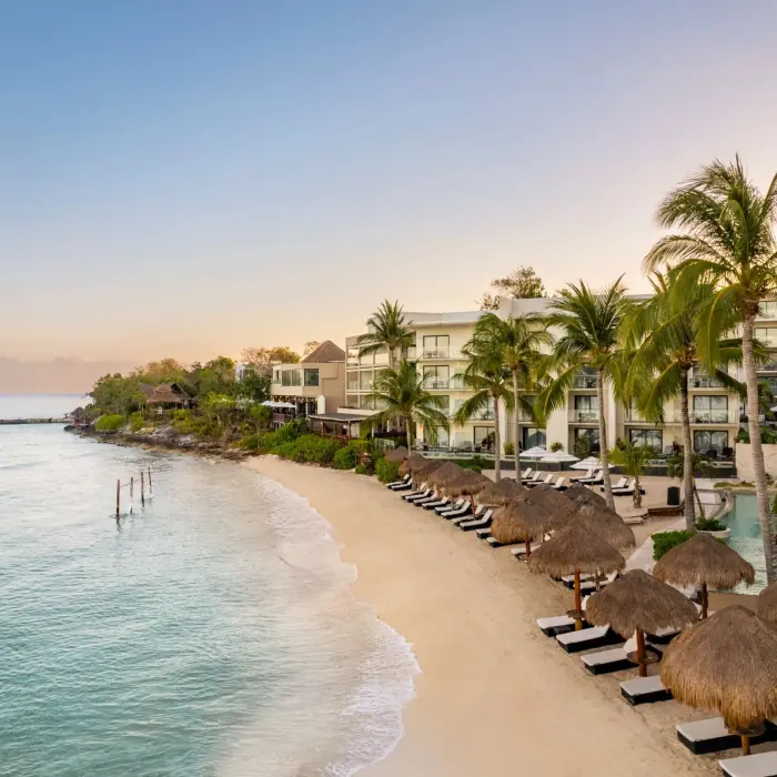 Beachfront resort at sunrise with ocean hammocks, palapas, loungers, and modern suites overlooking calm waters.