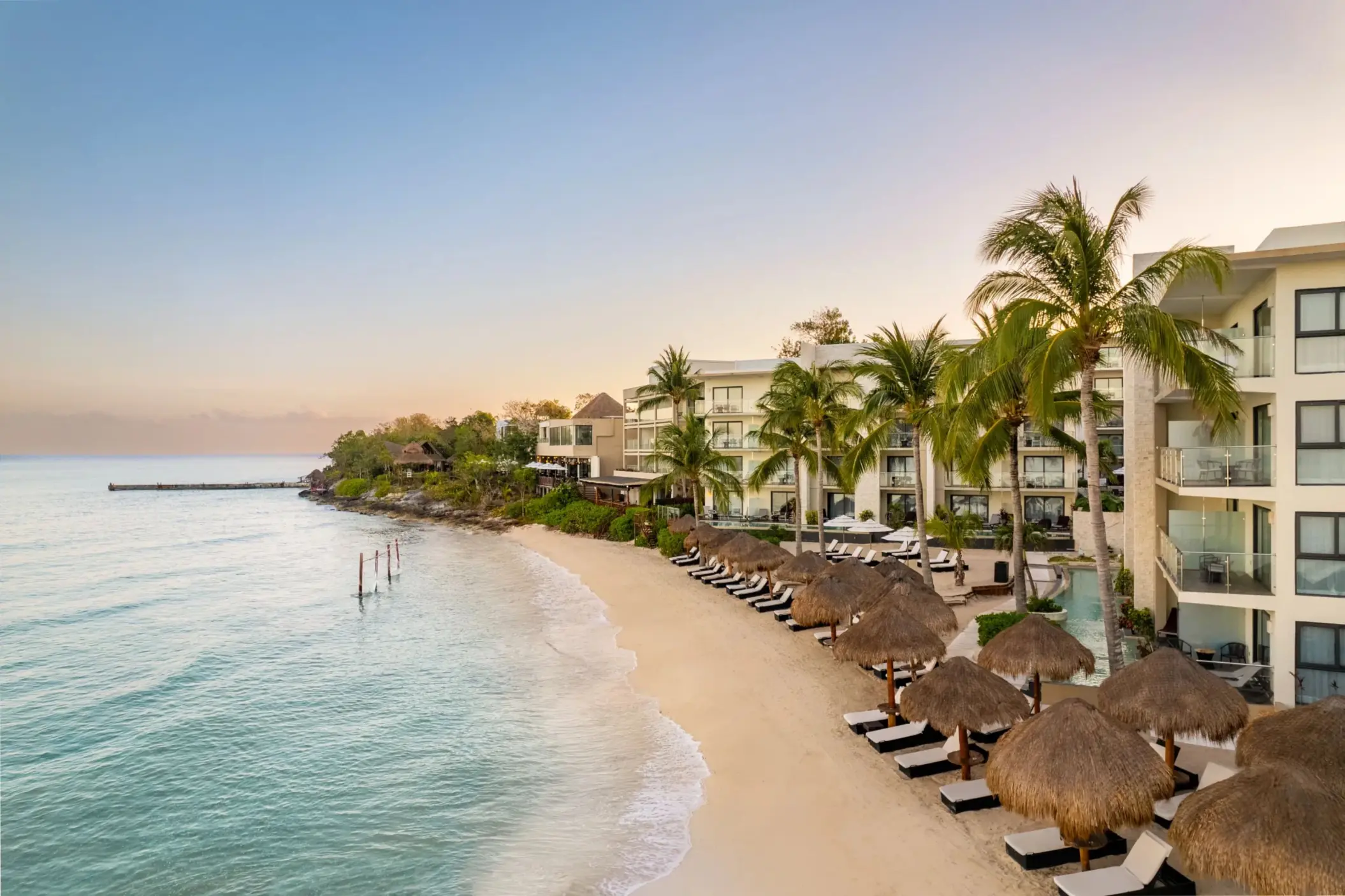 Beachfront resort at sunrise with ocean hammocks, palapas, loungers, and modern suites overlooking calm waters.