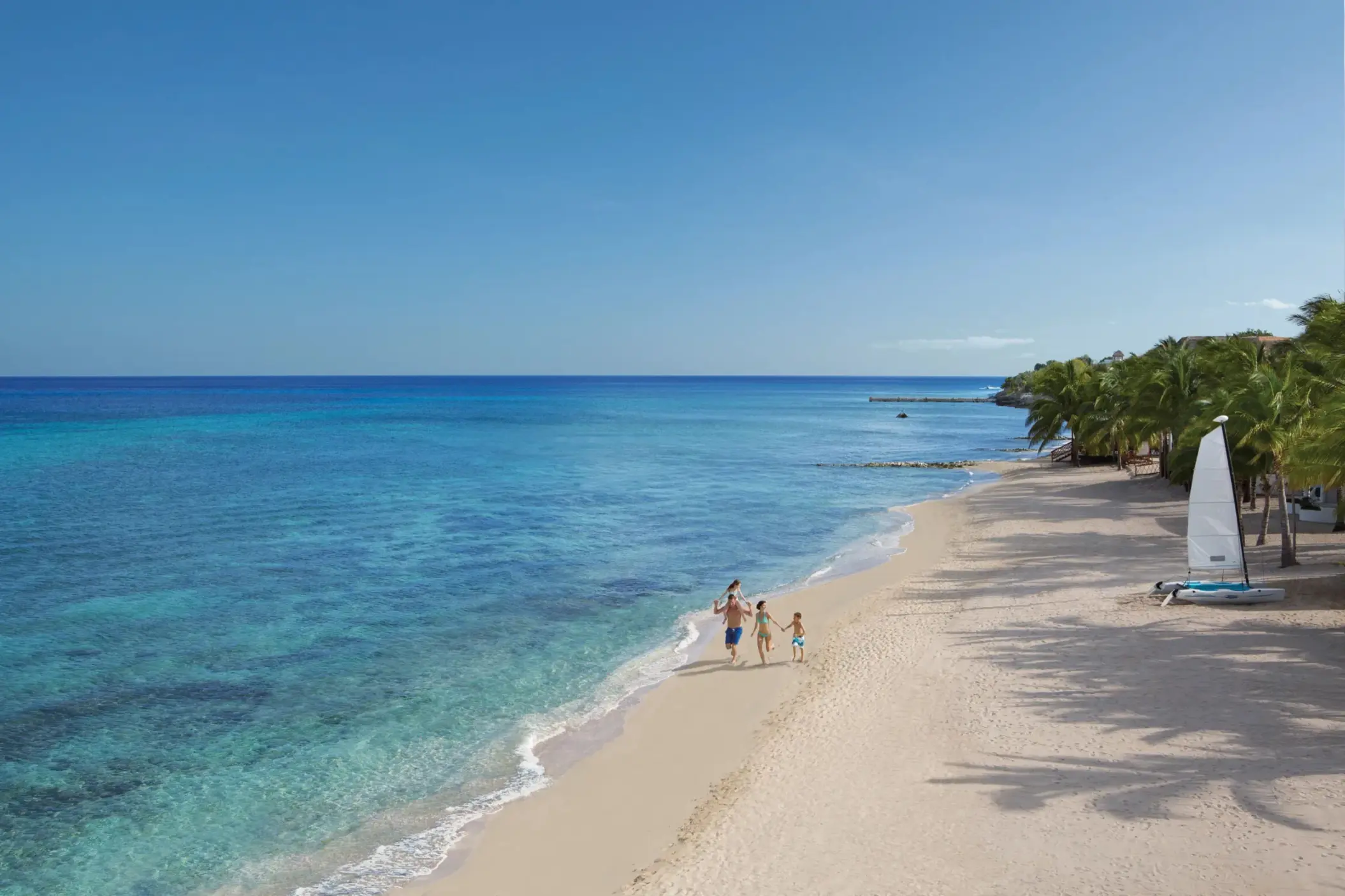 Family walking along the soft sandy beach beside crystal-clear waters and palm-shaded shoreline.