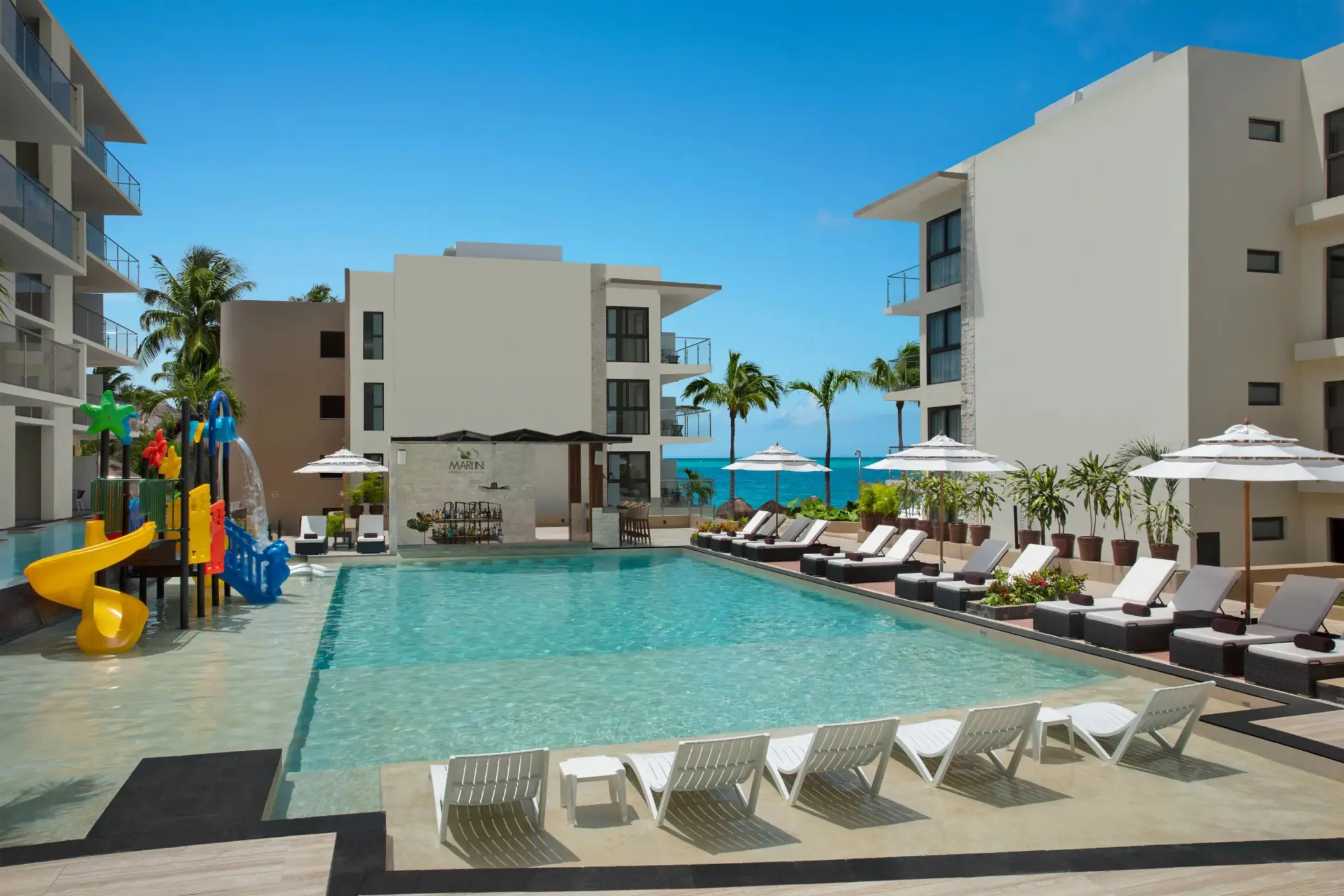 Main resort pool with loungers, umbrellas, splash zone, and Marlin swim-up bar under blue skies.