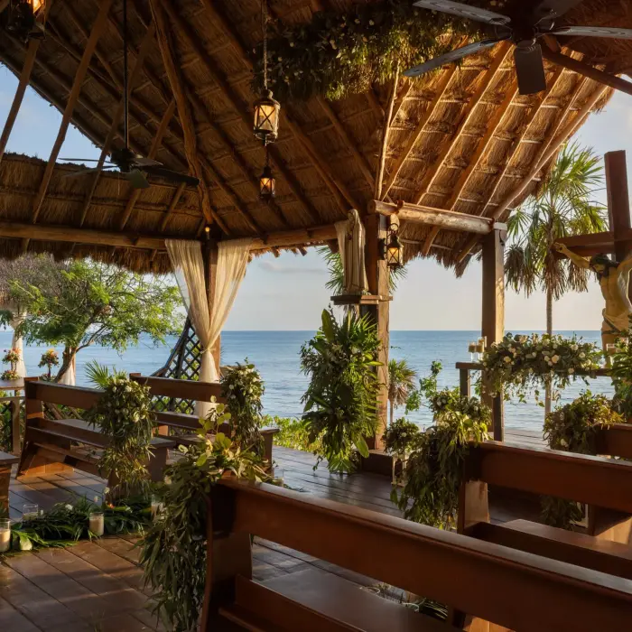 Oceanfront chapel setup with rustic wooden benches, tropical greenery, and religious statues under palapa roof.