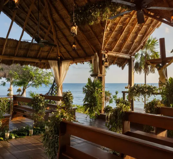 Oceanfront chapel setup with rustic wooden benches, tropical greenery, and religious statues under palapa roof.