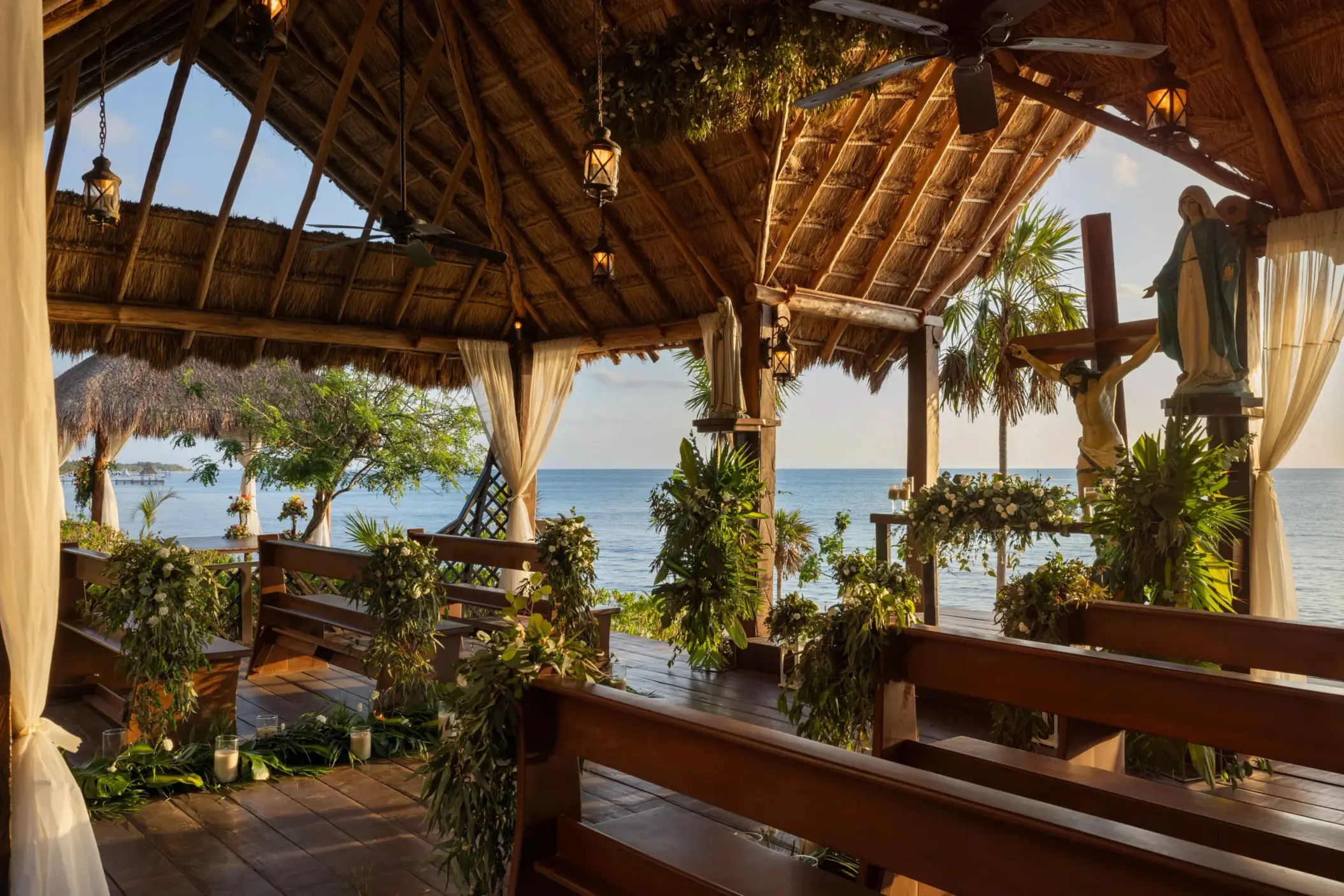 Oceanfront chapel setup with rustic wooden benches, tropical greenery, and religious statues under palapa roof.