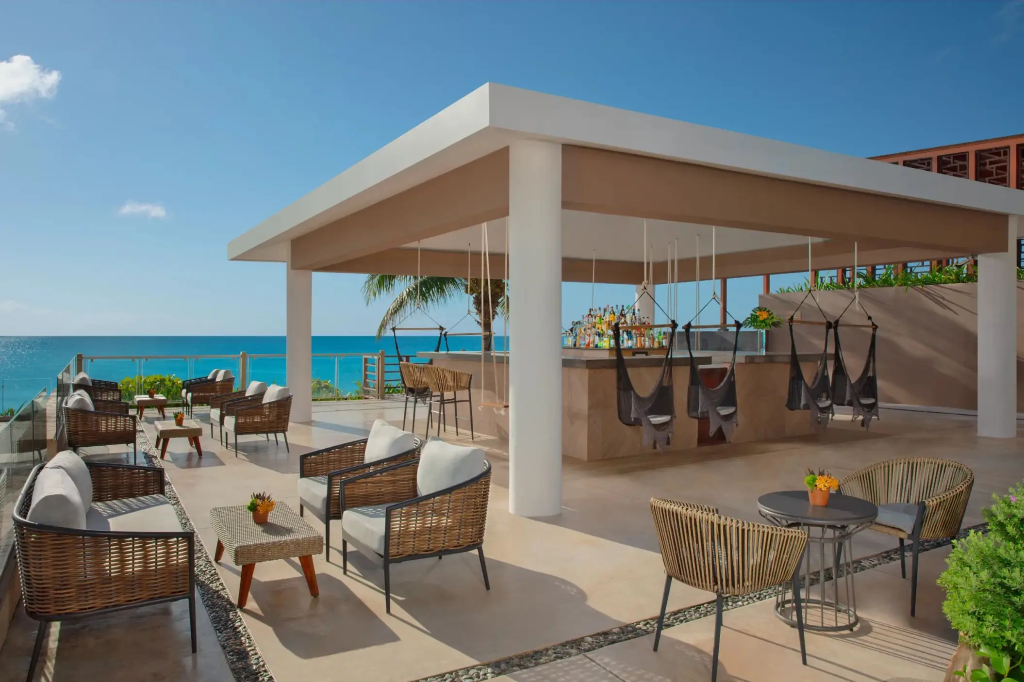 Modern beachfront rooftop bar with wicker furniture, hanging swing chairs, ocean views, and tropical setting