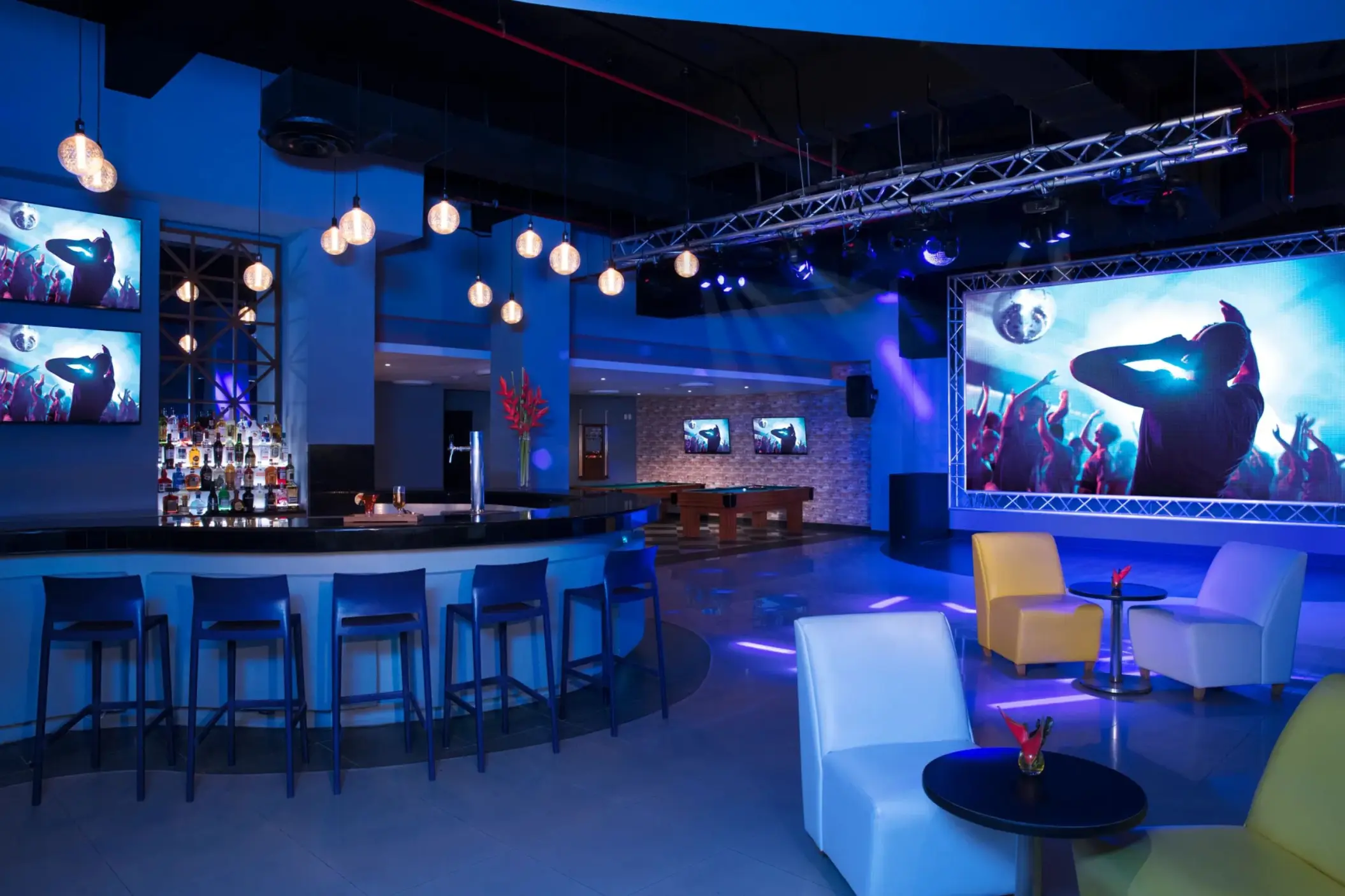 Contemporary nightclub interior with blue lighting, projection screens, bar seating, and modern lounge furniture