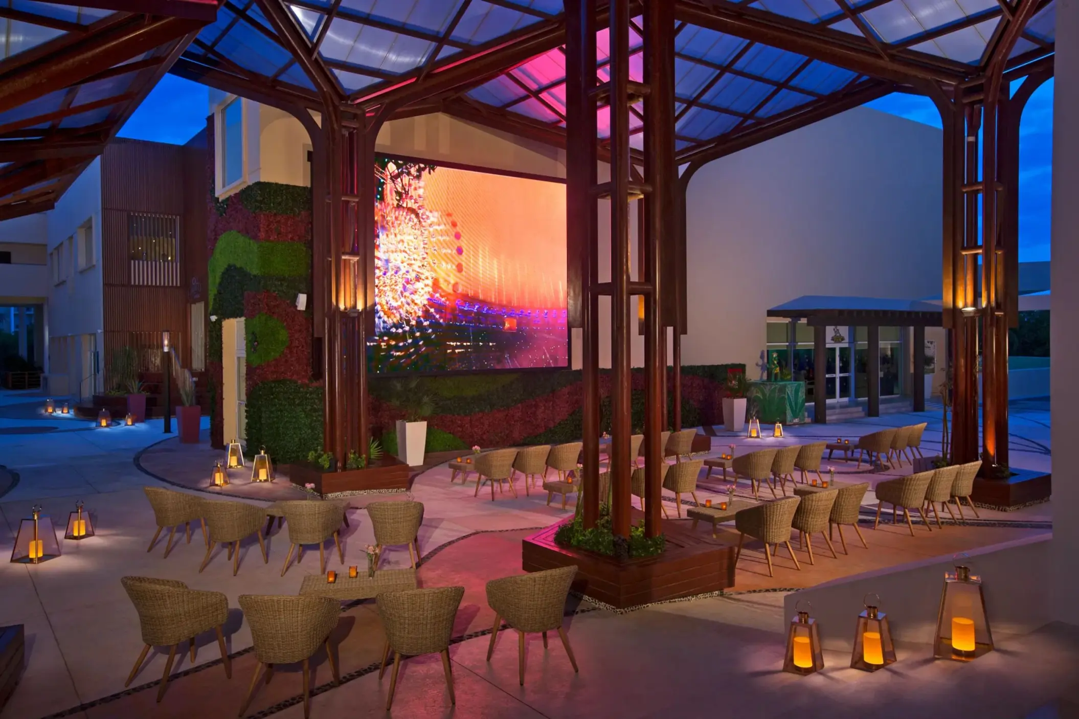 Modern outdoor bar pavilion with glass roof, wicker seating, large screen, and ambient evening lighting.