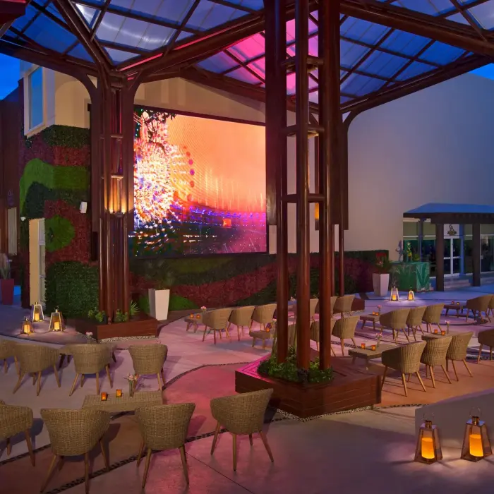 Modern outdoor bar pavilion with glass roof, wicker seating, large screen, and ambient evening lighting.