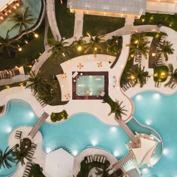 Close-up aerial of resort pool area with curved bridges, palm trees, and central glass-walled room over water.
