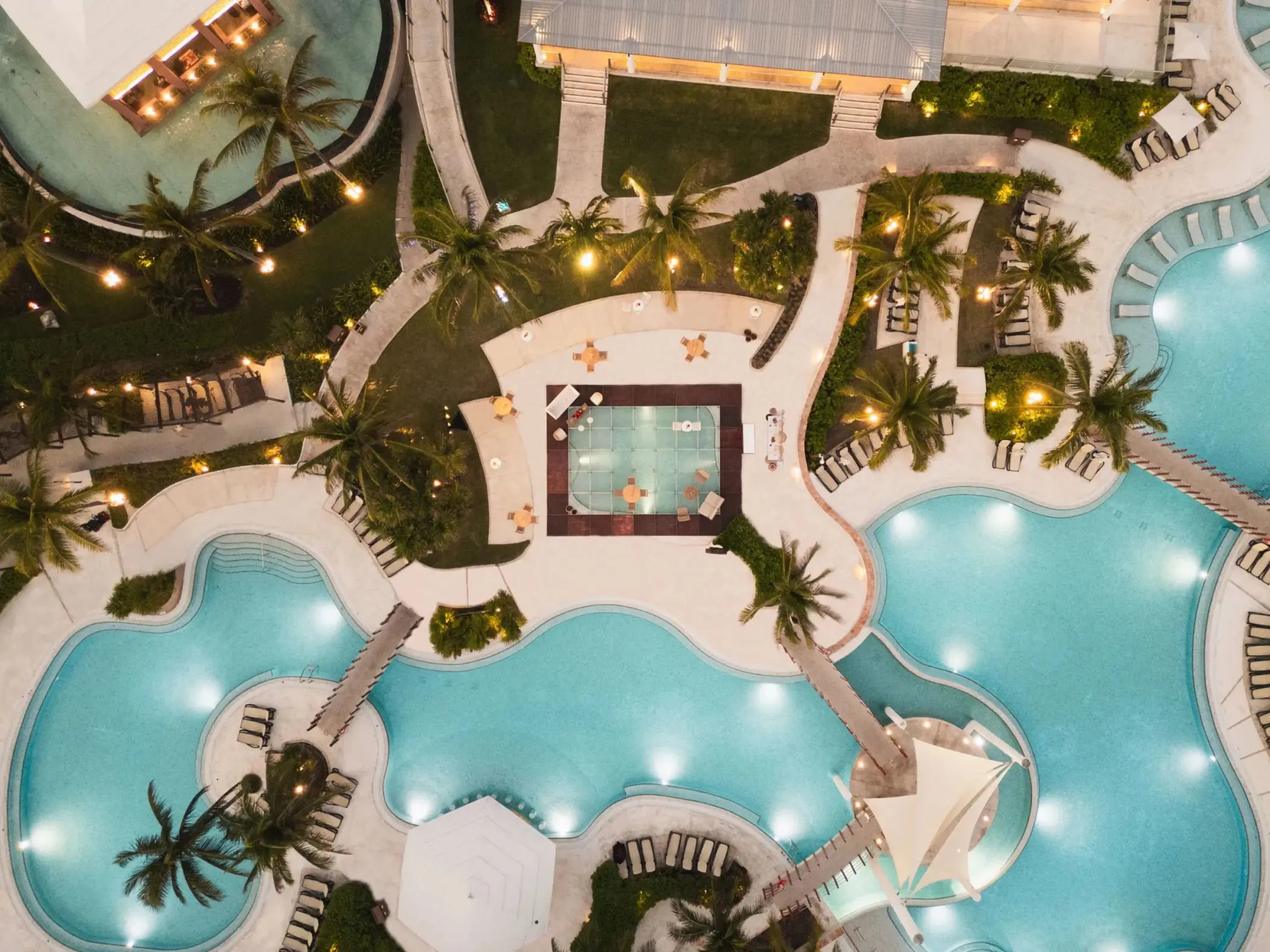 Close-up aerial of resort pool area with curved bridges, palm trees, and central glass-walled room over water.