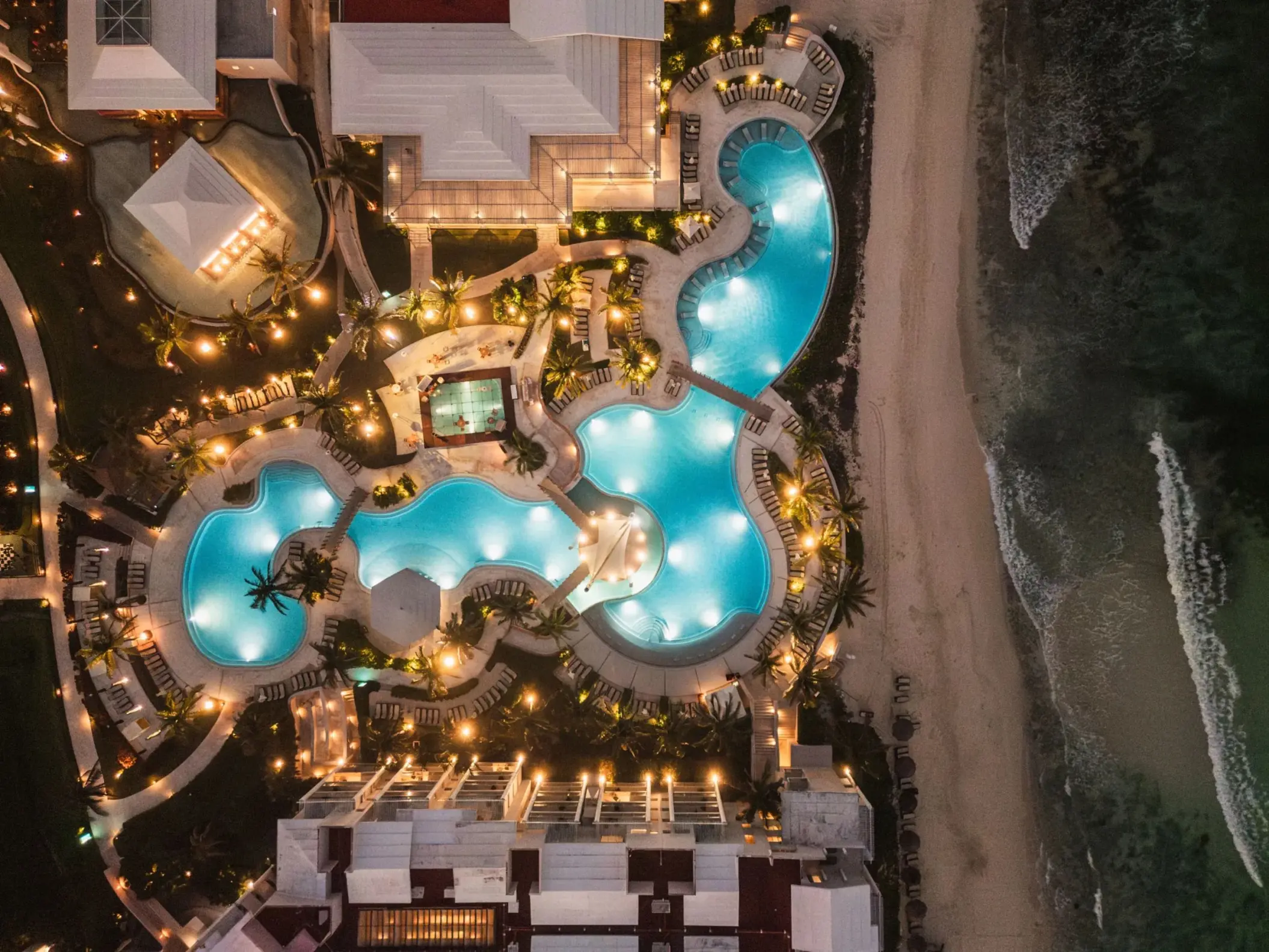 Nighttime aerial view of resort pools and beachfront with glowing lights highlighting the elegant layout.