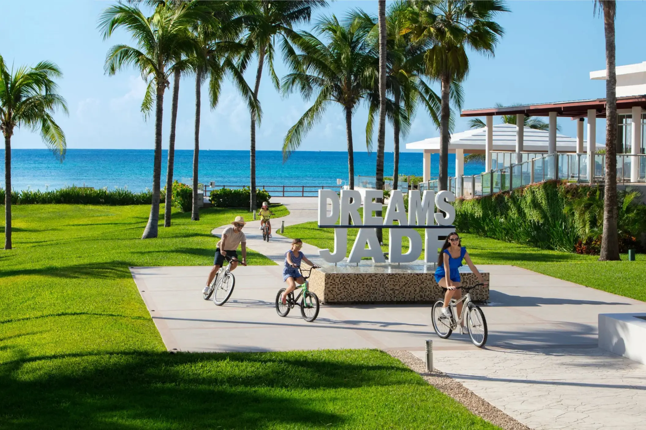 Family riding bikes near the Dreams Jade Resort sign with palm trees and ocean in the background.