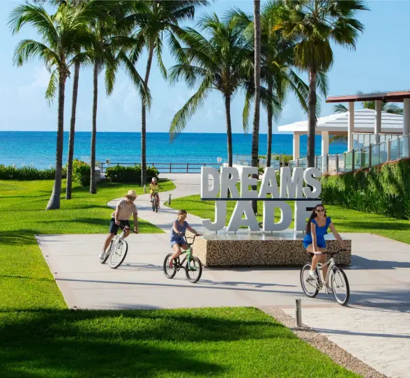 Family riding bikes near the Dreams Jade Resort sign with palm trees and ocean in the background.