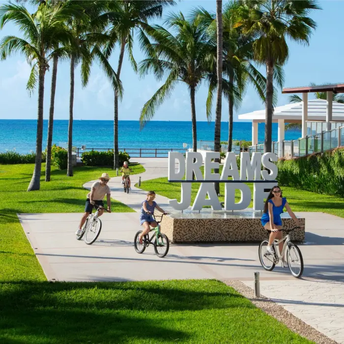 Family riding bikes near the Dreams Jade Resort sign with palm trees and ocean in the background.
