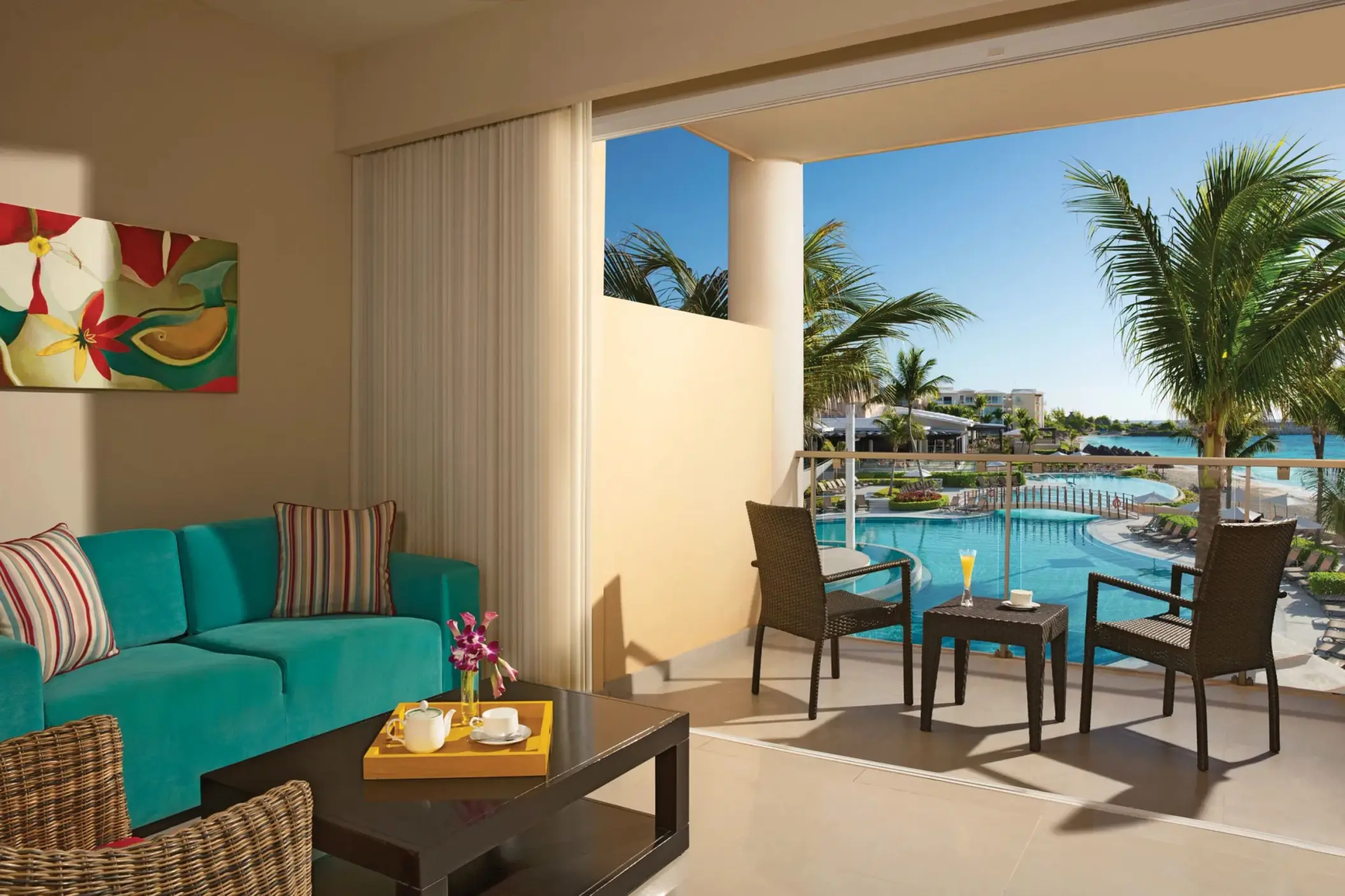 Junior suite balcony view with teal sofa and tropical drinks overlooking the pool and resort landscape.
