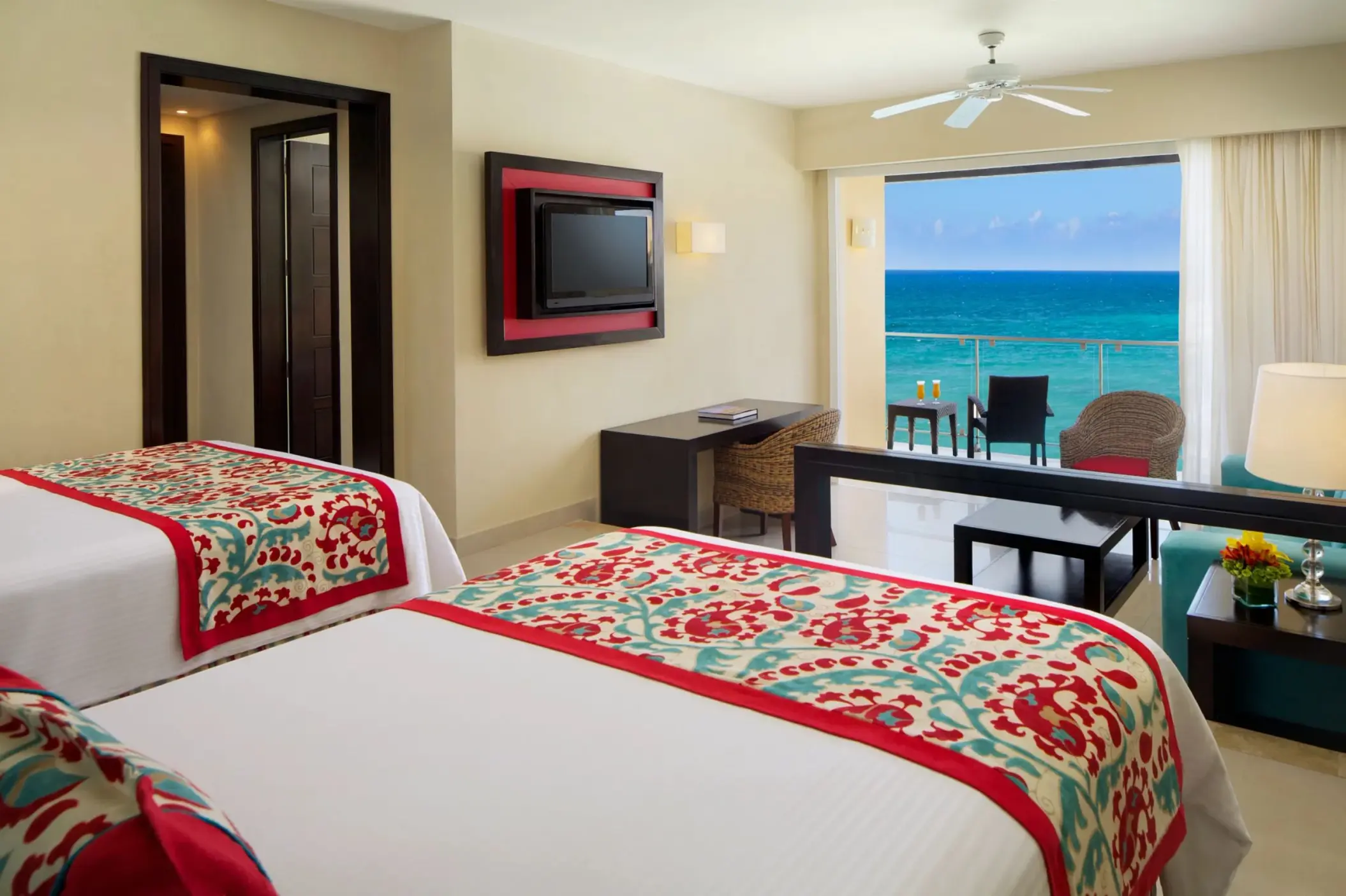 Spacious double bed suite with oceanfront balcony, wicker seating, and vibrant red and teal decor.