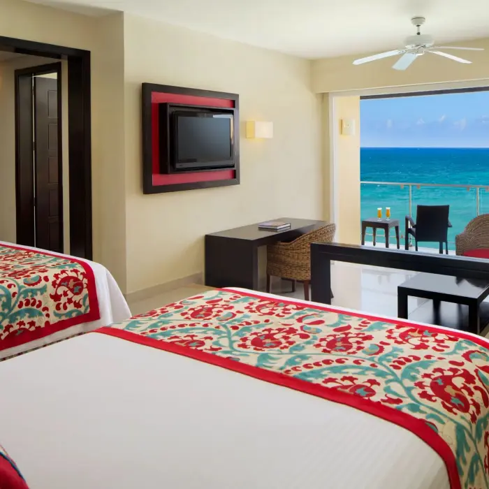 Spacious double bed suite with oceanfront balcony, wicker seating, and vibrant red and teal decor.