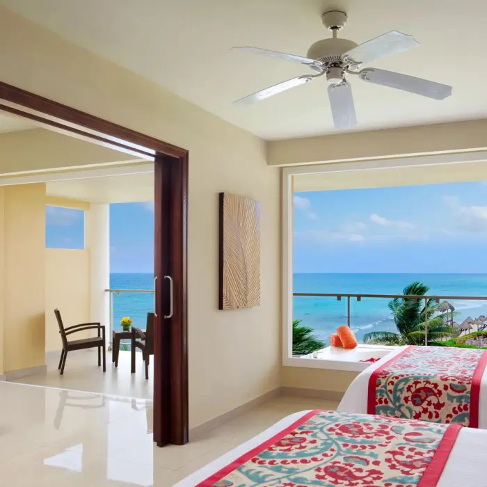 Bright oceanview suite with two beds, sitting area, and panoramic windows overlooking turquoise waters.