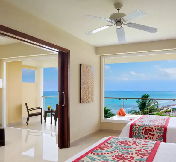 Bright oceanview suite with two beds, sitting area, and panoramic windows overlooking turquoise waters.