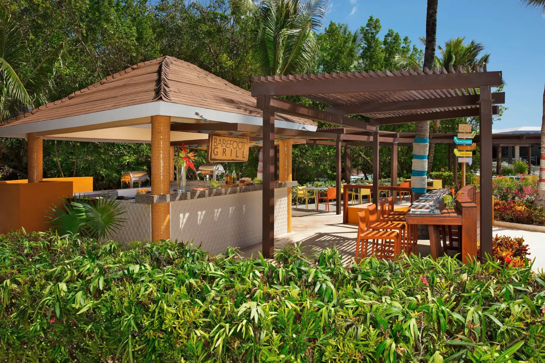 Colorful poolside grill station surrounded by tropical greenery and shaded seating areas.