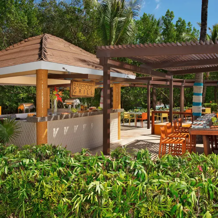 Colorful poolside grill station surrounded by tropical greenery and shaded seating areas.