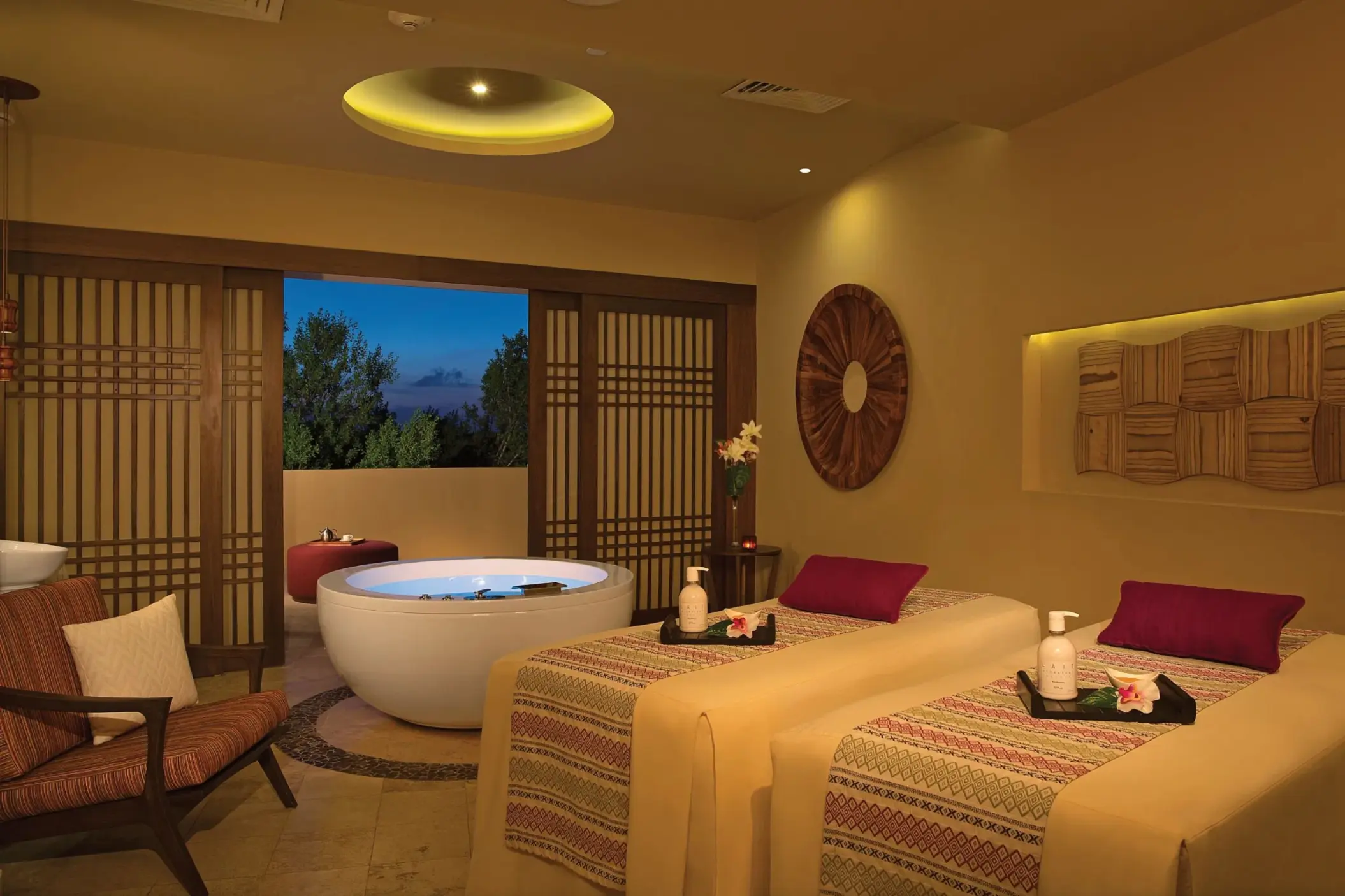 Elegant spa cabin with twin massage beds, wooden decor, and a private outdoor tub set against evening skies.