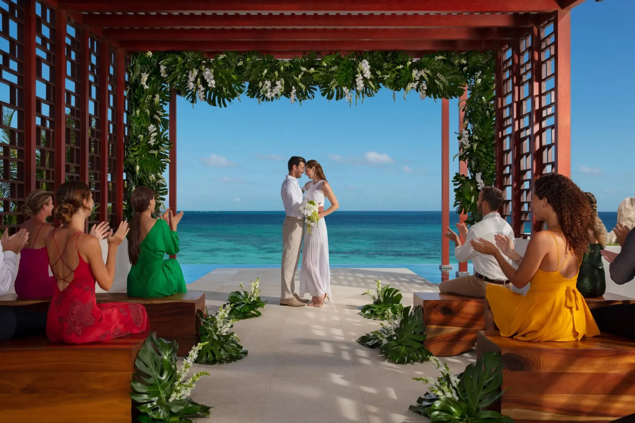 Bride and groom exchange vows under a floral pergola facing the ocean, surrounded by seated guests applauding.