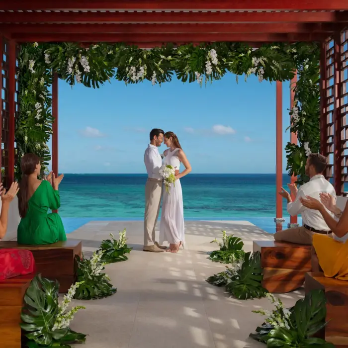 Bride and groom exchange vows under a floral pergola facing the ocean, surrounded by seated guests applauding.
