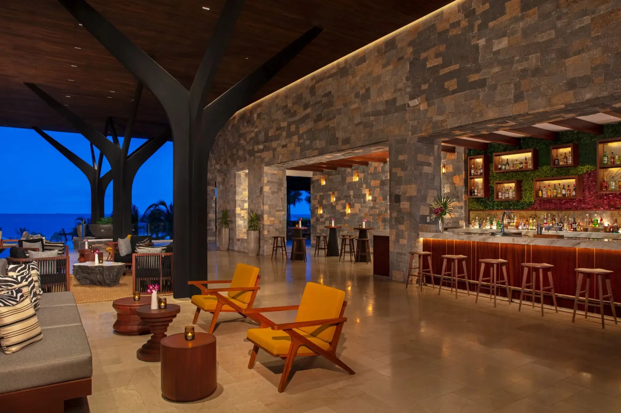 Elegant open-air bar with stone walls, warm lighting, and ocean views at night in Rendezvous Lounge.