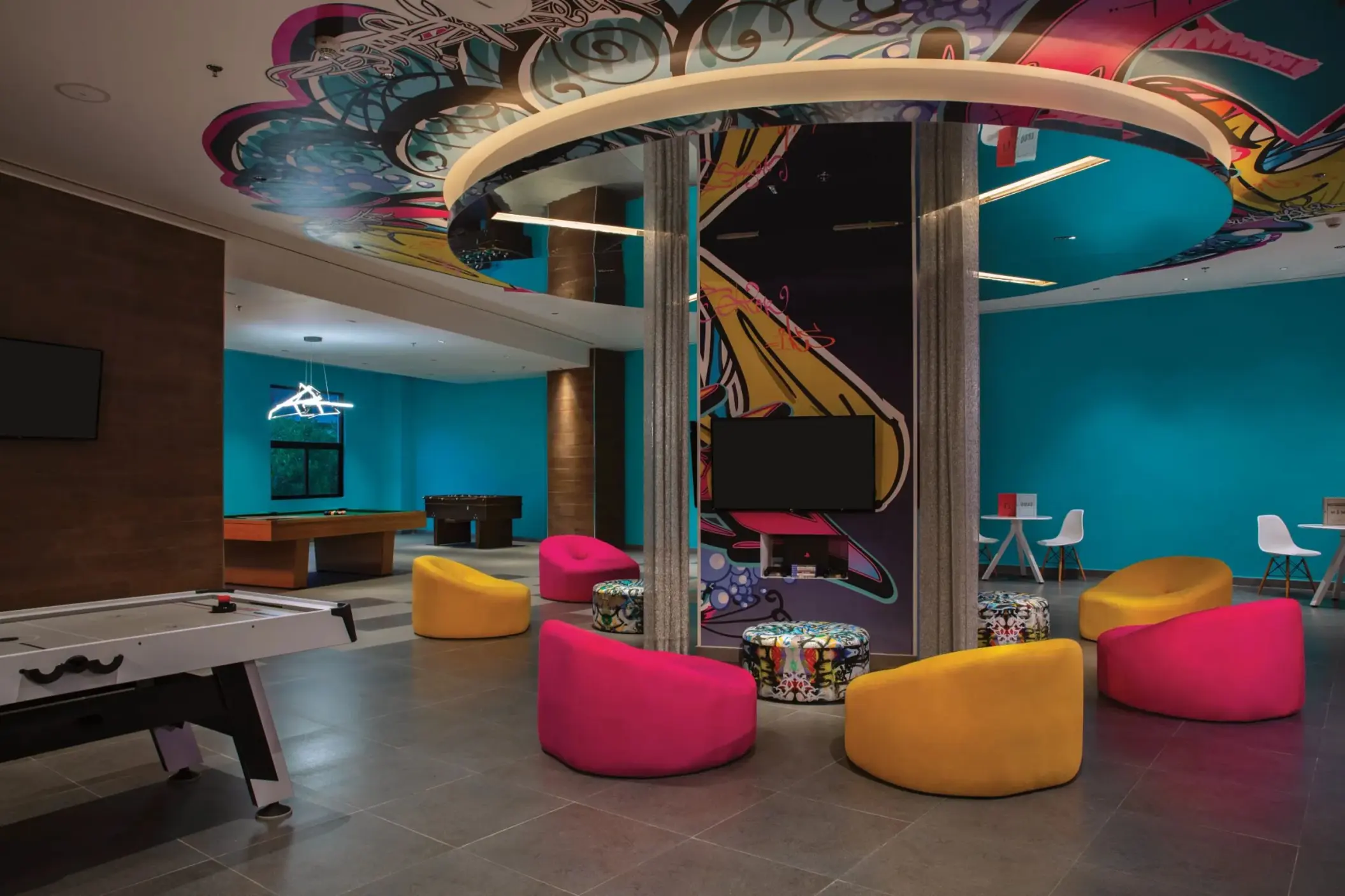 Vibrant teen club with colorful seating, arcade games, and bold graffiti art on the ceiling and central pillar.