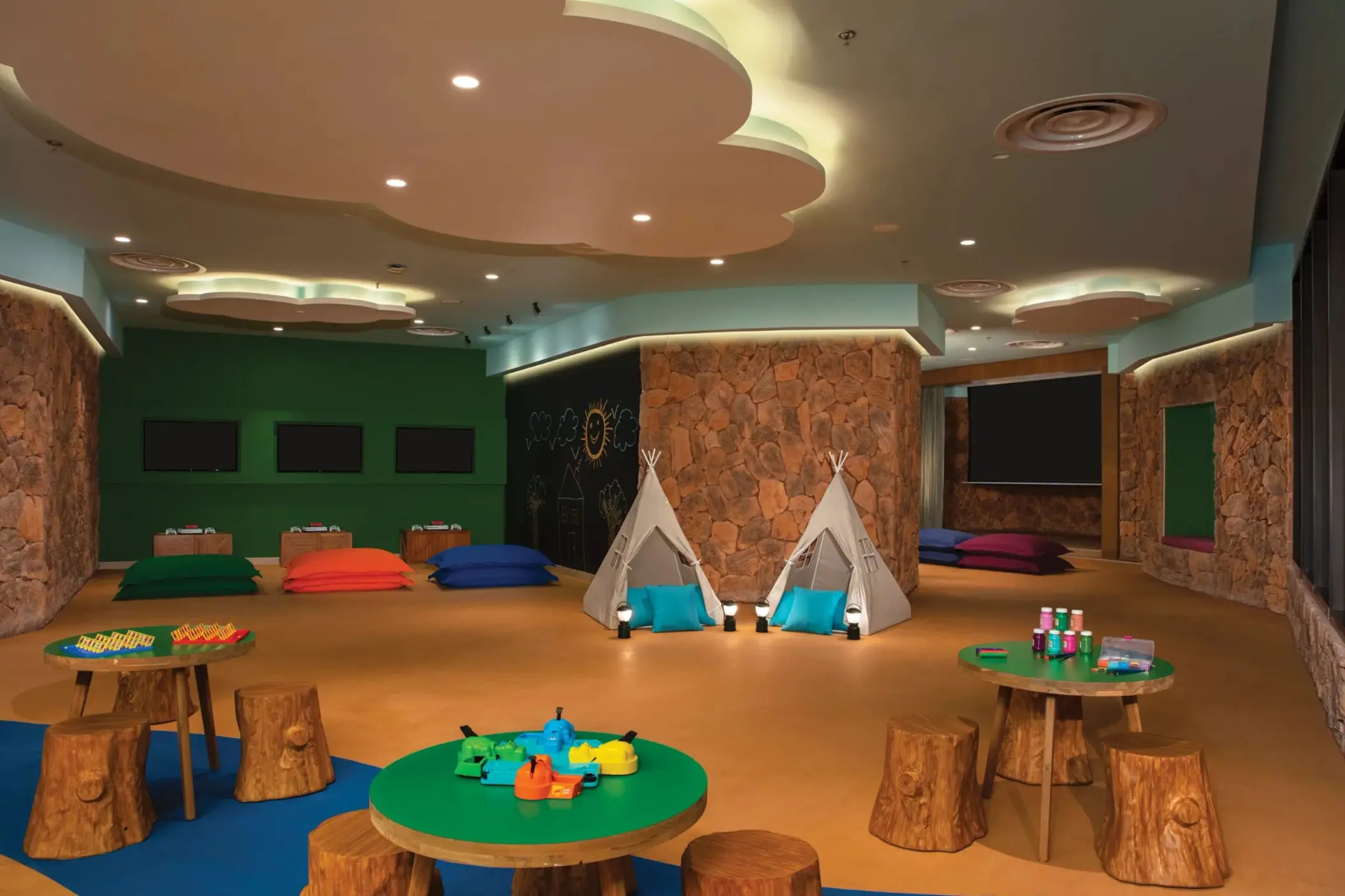 Kids’ indoor club with stone walls, teepees, game tables, and colorful floor cushions.