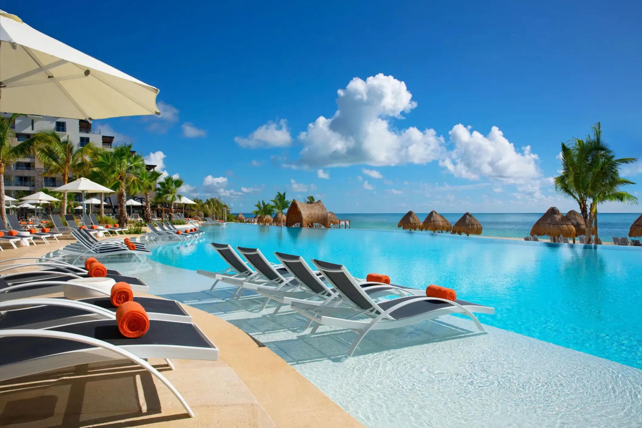 Infinity pool lined with loungers and umbrellas, overlooking the ocean with palapas and tropical palms.