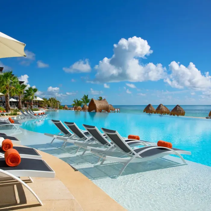 Infinity pool lined with loungers and umbrellas, overlooking the ocean with palapas and tropical palms.