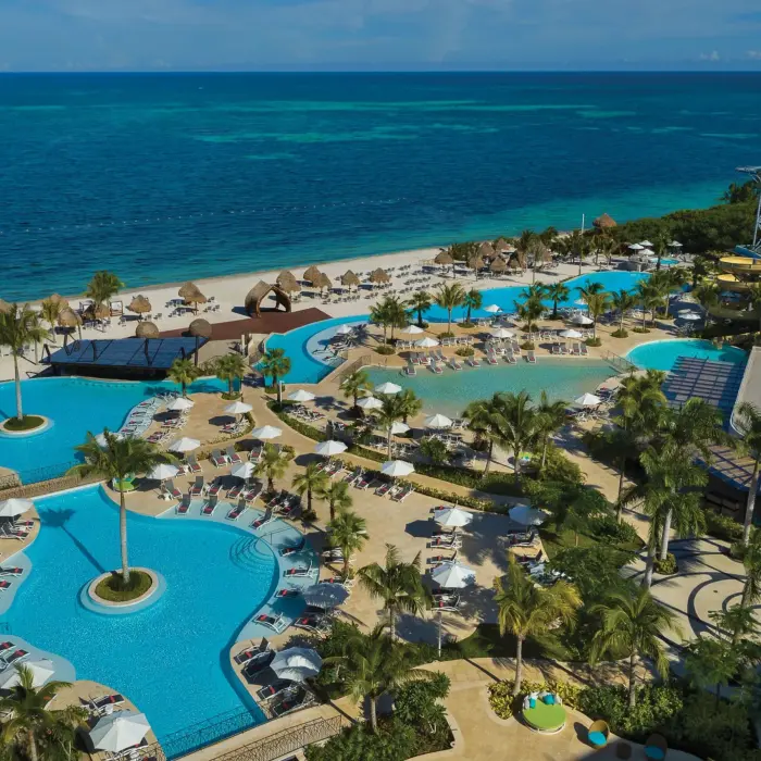 Aerial view of resort pools by the beach, featuring loungers, palm trees, and water slides under bright blue sky.