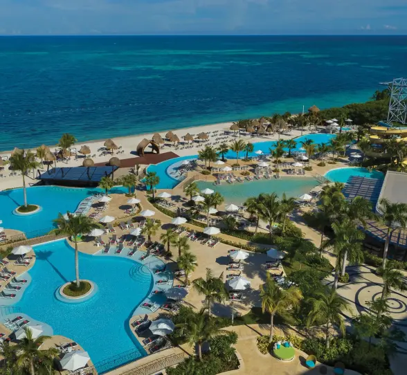 Aerial view of resort pools by the beach, featuring loungers, palm trees, and water slides under bright blue sky.