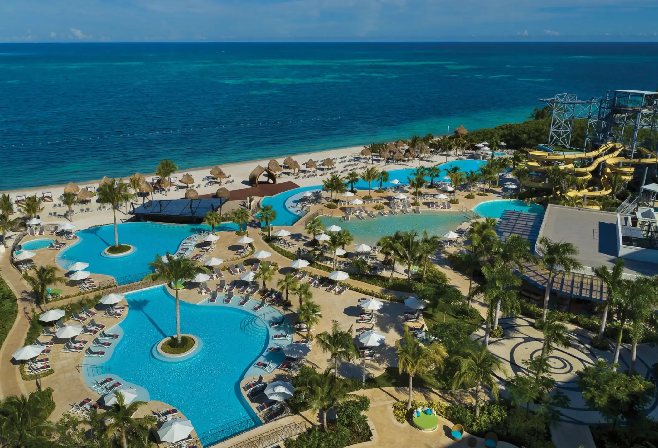 Aerial view of resort pools by the beach, featuring loungers, palm trees, and water slides under bright blue sky.