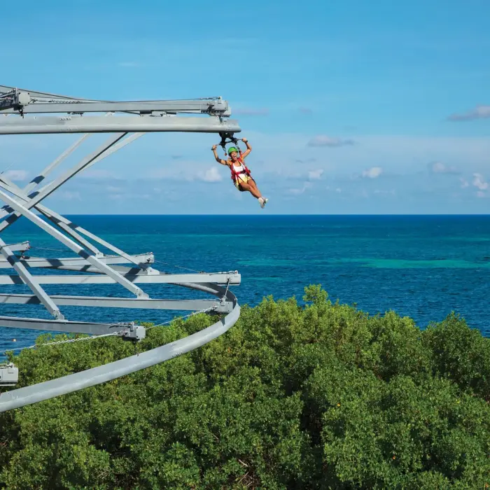 Guest riding a rollglider zipline over lush greenery with panoramic views of the Caribbean Sea.