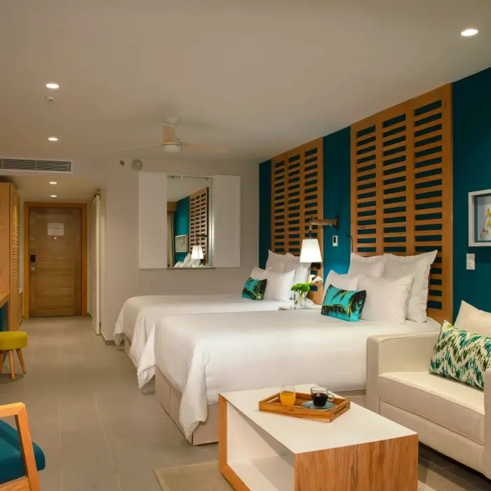 Modern junior suite with two beds, teal accents, wood furnishings, and cozy sitting area with wall decor.
