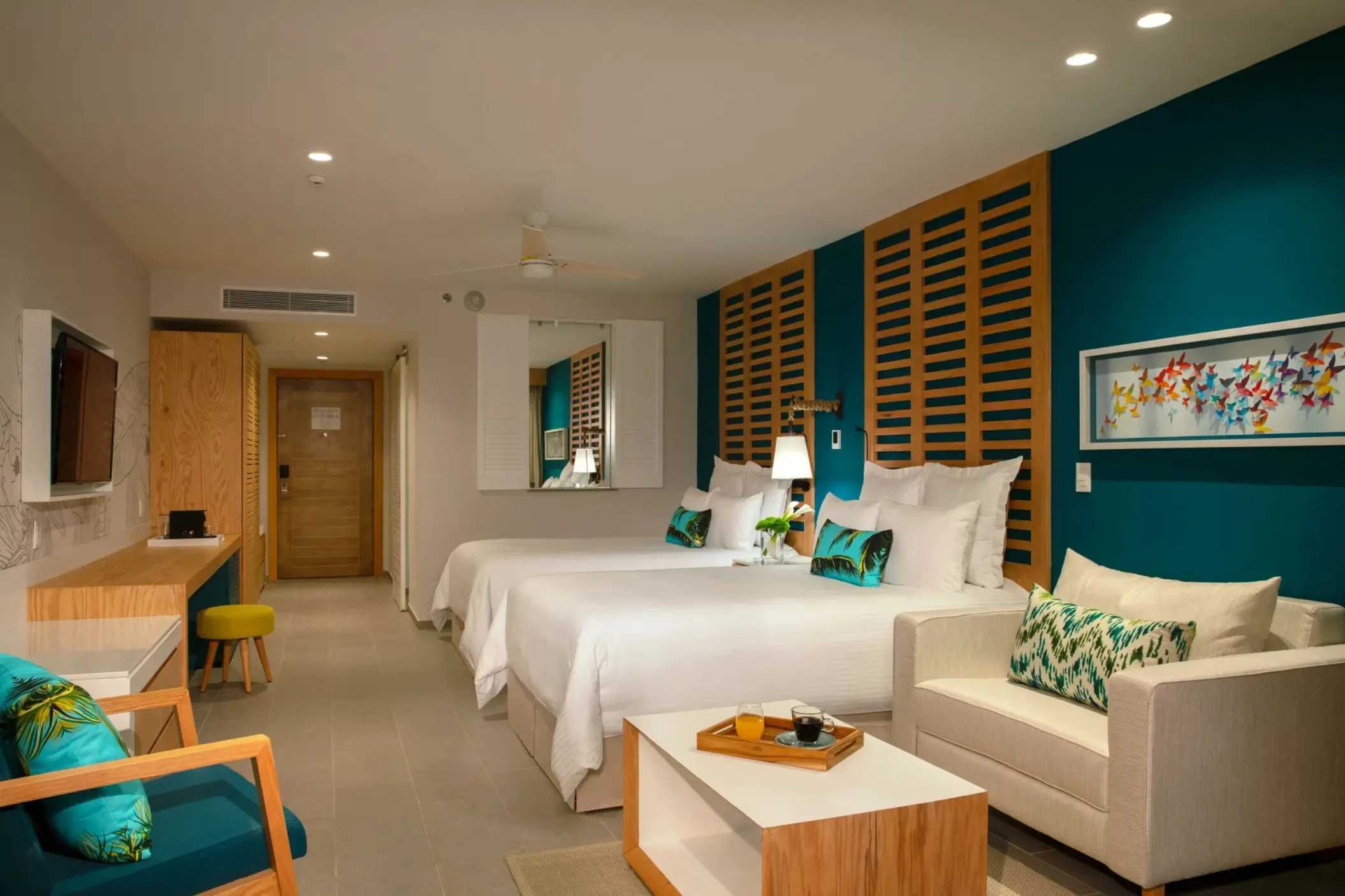 Modern junior suite with two beds, teal accents, wood furnishings, and cozy sitting area with wall decor.