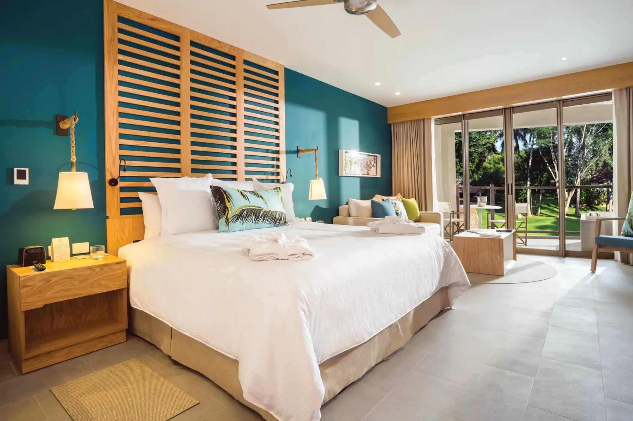 Spacious guestroom with teal accent wall, king bed, seating area and balcony view of garden greenery and palm trees.