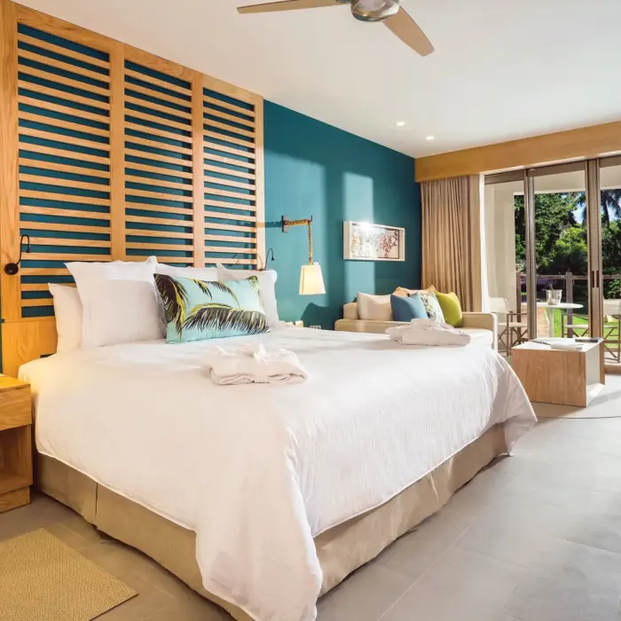 Spacious guestroom with teal accent wall, king bed, seating area and balcony view of garden greenery and palm trees.