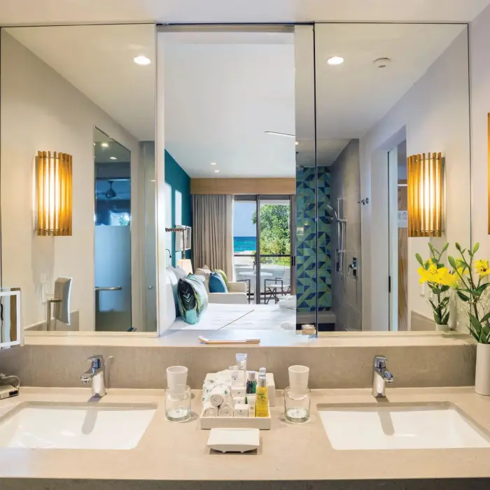 Modern double-sink vanity with warm lighting, view into bedroom and ocean beyond through open bathroom doorway.