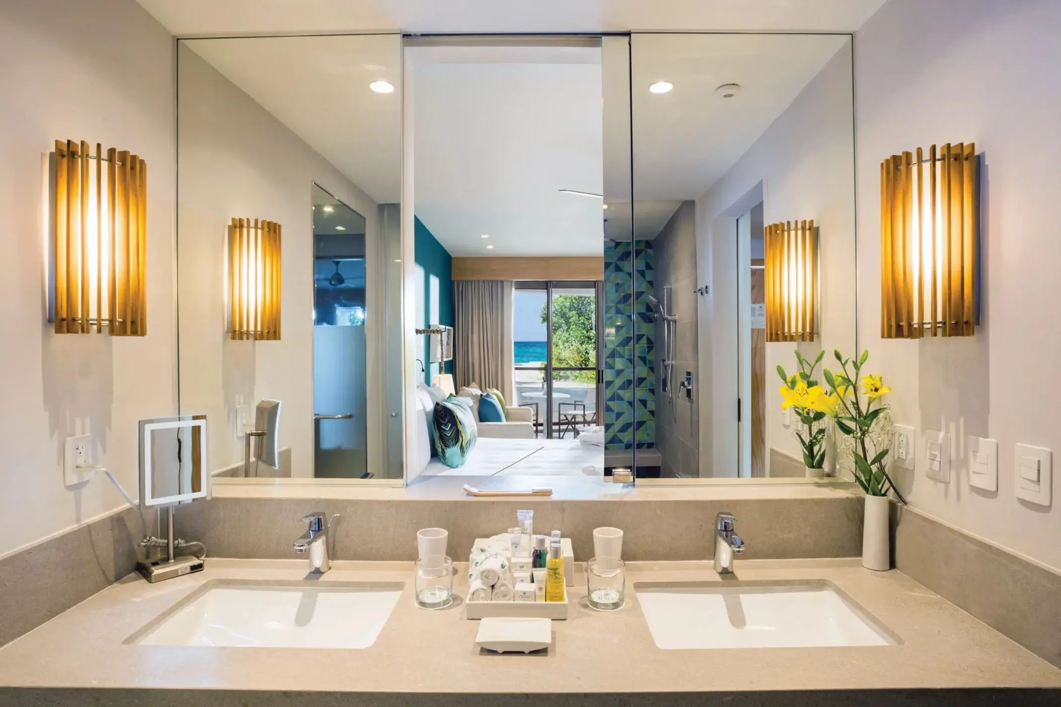 Modern double-sink vanity with warm lighting, view into bedroom and ocean beyond through open bathroom doorway.