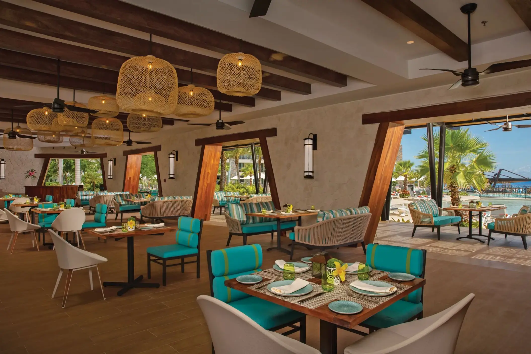 Stylish open-air restaurant with turquoise seating, wooden beams, and poolside views surrounded by palm trees.