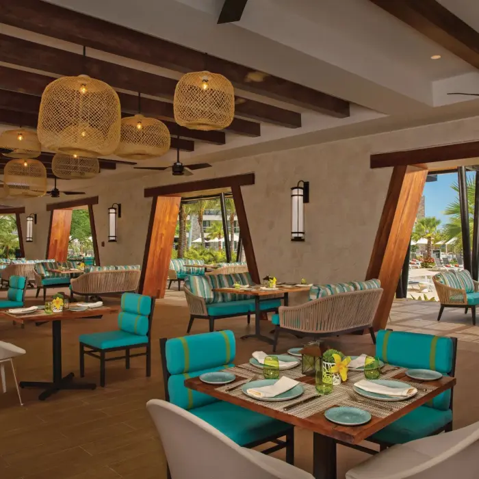 Stylish open-air restaurant with turquoise seating, wooden beams, and poolside views surrounded by palm trees.
